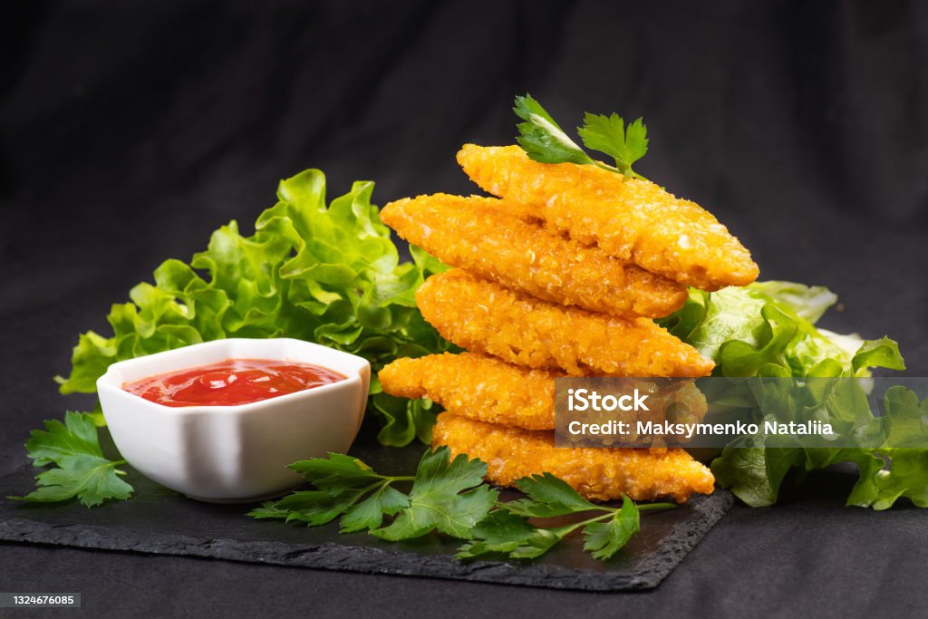 Fast homemade food.Chicken breaded nuggets on a black kitchen plate with ketchup and basil leaves, lettuce and tomatoes on a wooden background.